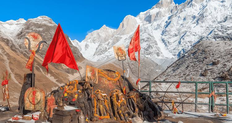 Sanctuaire de roche sacrée orné de drapeaux rouges et d'offrandes adossé à des pics enneigés escarpés.