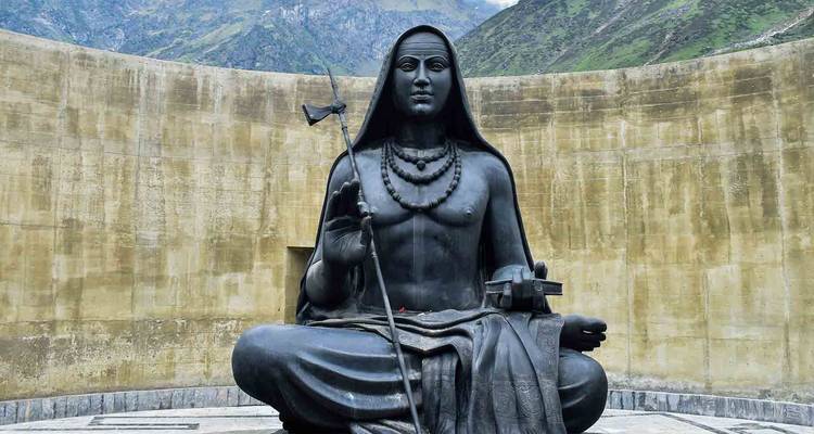Grande statue d'Adi Shankaracharya assis en méditation contre un mur de pierre circulaire avec des montagnes en arrière-plan.