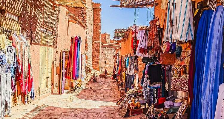 Narrow sunlit alley of earthen buildings showcasing colorful fabrics and handicrafts in Aït Benhaddou.