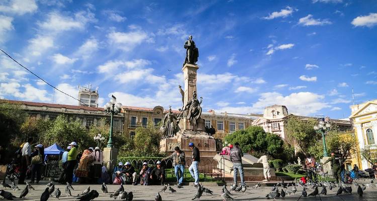 Place animée avec statue historique, habitants, vendeurs et volées de pigeons sur fond de ciel éclatant.
