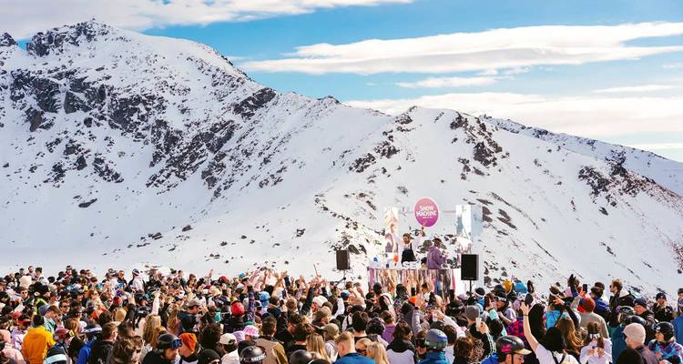 Große Menschenmenge genießt eine Musikveranstaltung im Freien auf einem verschneiten Berghang mit Gipfeln im Hintergrund.