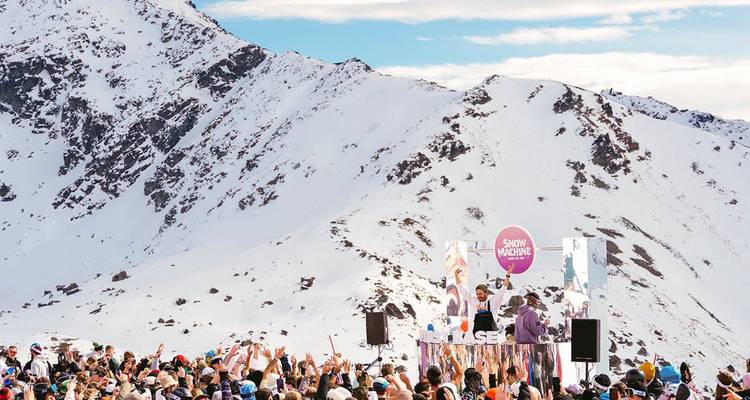 A lively crowd gathers for an outdoor DJ set on a snowy mountain ridge with dramatic peaks behind.
