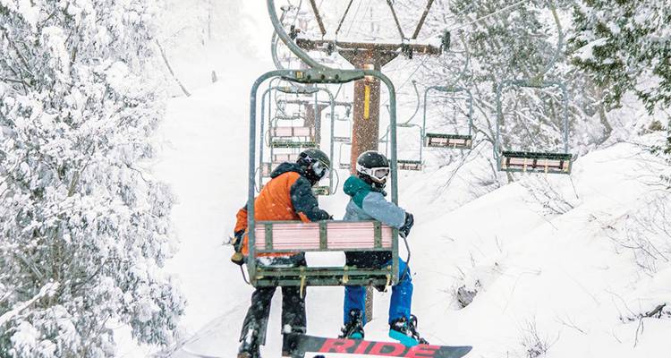 Dos snowboarders van en un telesilla nevado a través de un denso bosque invernal.