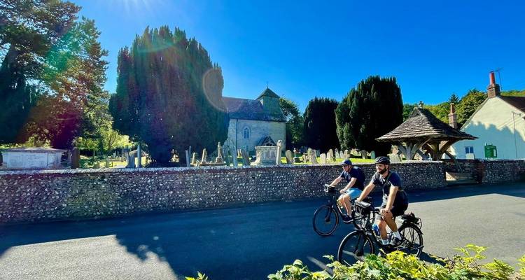 Deux cyclistes passent devant un vieux cimetière en pierre sous un soleil éclatant et un ciel bleu vibrant.