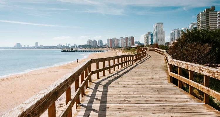 Wooden boardwalk running along sandy city beach with skyline of Punta del Este in background.
