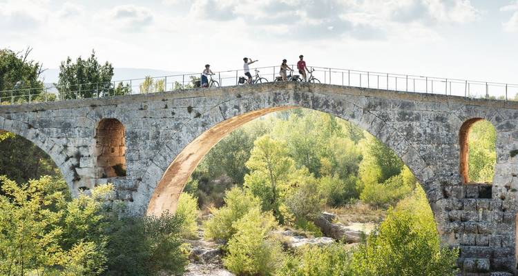 Pequeño grupo de ciclistas hace una pausa en la cima de un puente de piedra histórico rodeado de bosque verde.