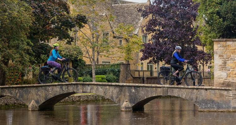 Zwei Radfahrer überqueren eine kleine Steinbrücke über einen ruhigen Bach in einem grünen Dorf in den Cotswolds