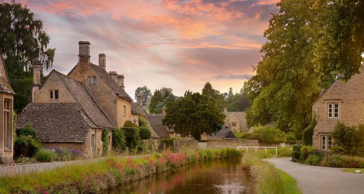 Typische Cotswolds-Steinhäuser säumen einen ruhigen Kanal bei Sonnenuntergang mit pastellfarbenen Wolken, die sich im Wasser spiegeln