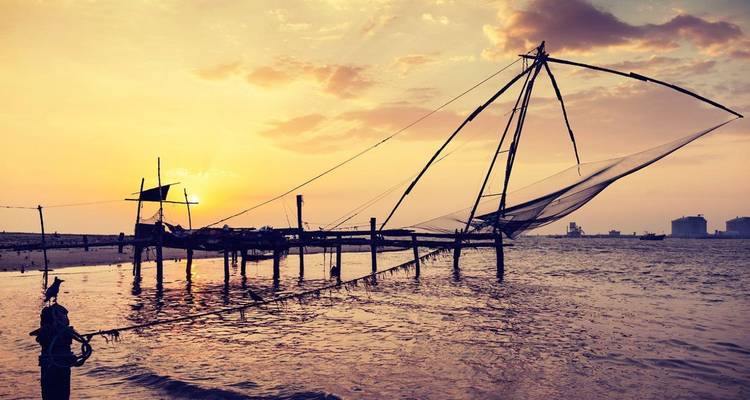 Traditionelles chinesisches Fischernetz als Silhouette vor einem orangefarbenen Sonnenuntergang über den Backwaters von Kerala.