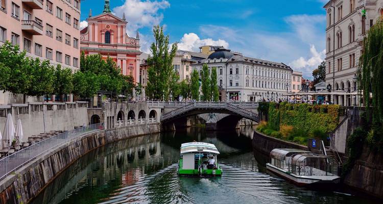 Un petit bateau navigue sous des ponts historiques le long de la charmante rivière de Ljubljana bordée de bâtiments aux couleurs pastel.