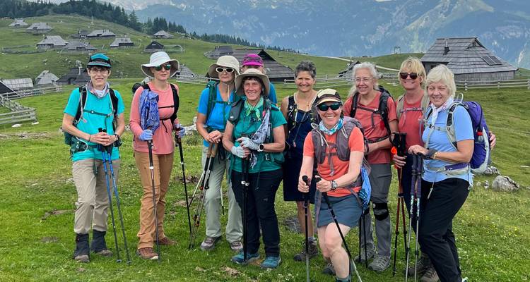 Un groupe joyeux de randonneuses pose avec des bâtons de randonnée sur un plateau alpin herbeux parsemé de cabanes en bois.