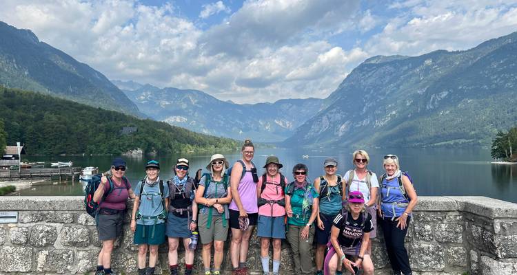 Grand groupe de randonneurs souriants sur une terrasse de pierre surplombant un lac serein et une chaîne de montagnes.