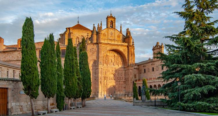 Fachada soleada de un gran monasterio de piedra arenisca con altos cipreses bordeando la plaza.