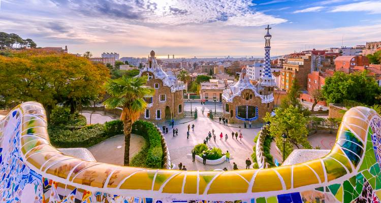 Colourful mosaic balustrade overlooking the central terrace of Park Güell with visitors scattered across the plaza and Barcelona skyline beyond.