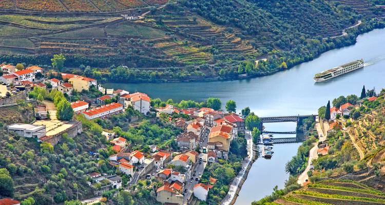 Luftaufnahme von terrassenförmigen Weinbergen und einer kleinen Stadt entlang des sich windenden Douro-Flusses mit einem Kreuzfahrtschiff.