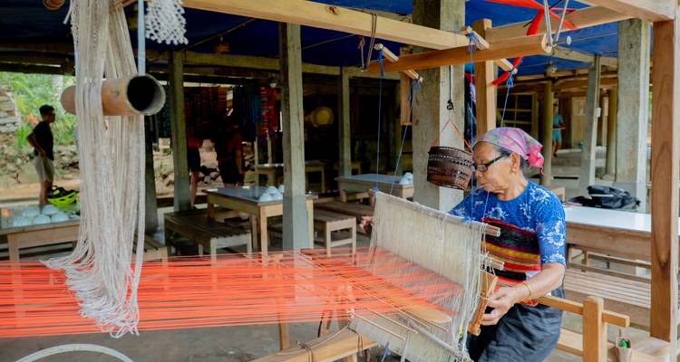 Tisserand âgé enfilant un fil vibrant à travers un métier à tisser en bois dans un atelier en plein air.