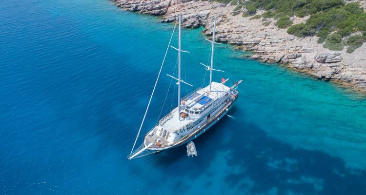 Aerial view of a lone gulet yacht anchored in crystal-clear turquoise water beside a rocky coast.