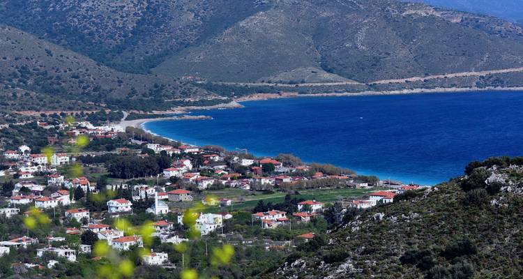 Elevated view of a coastal village with red-roofed houses nestled between green hills and a blue bay.