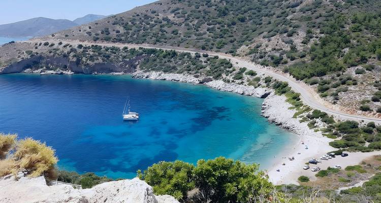 Curving coastal road overlooks a deep blue cove where a lone sailboat anchors in turquoise water.