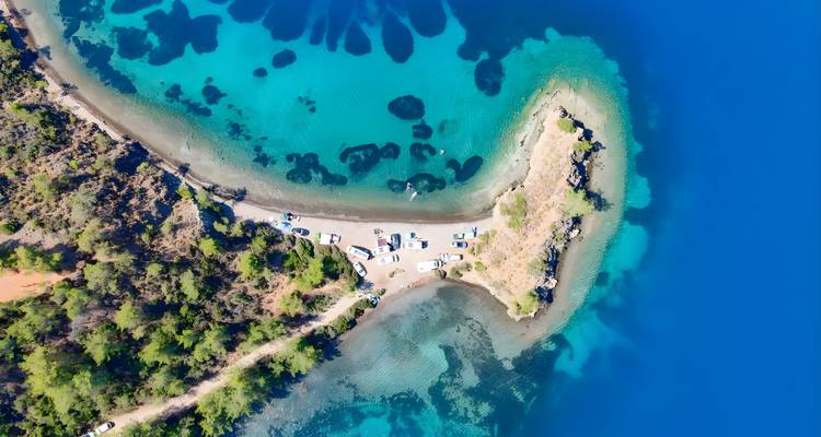 Top-down drone image of a narrow peninsula and parked cars surrounded by vibrant turquoise lagoons.