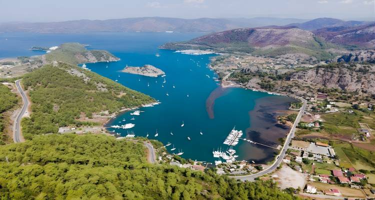 Aerial of a winding inlet filled with sailboats, surrounded by pine-covered hills and villages.