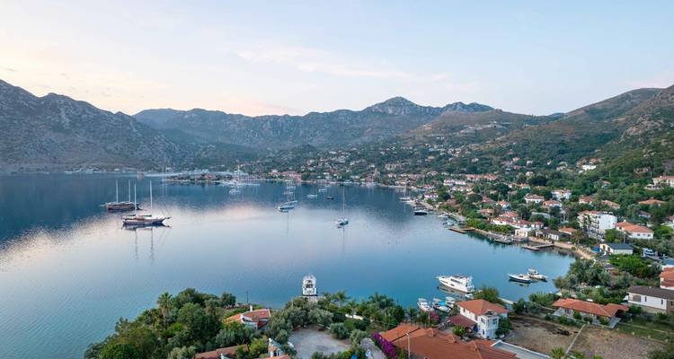 Calm bay town at dusk with moored yachts reflecting soft pastel sky, backed by rugged hills.
