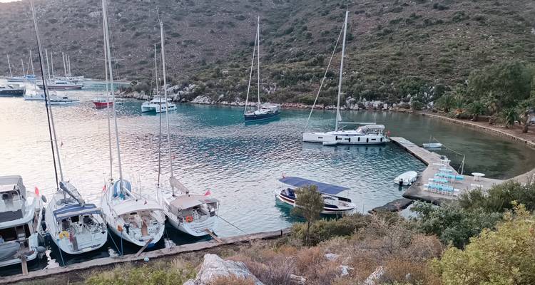 Small marina with sailboats tied to a stone quay in a quiet cove lined by scrub hills.