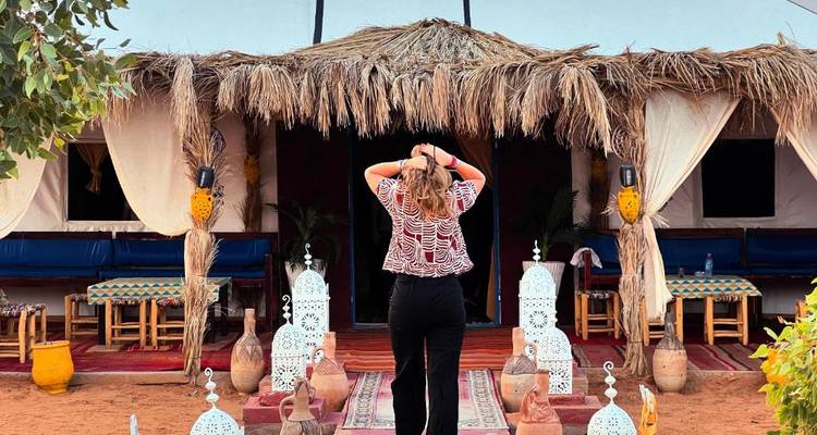 Une femme se tient à l'entrée d'un campement dans le désert décoré de lanternes, de tapis et d'auvents de chaume.