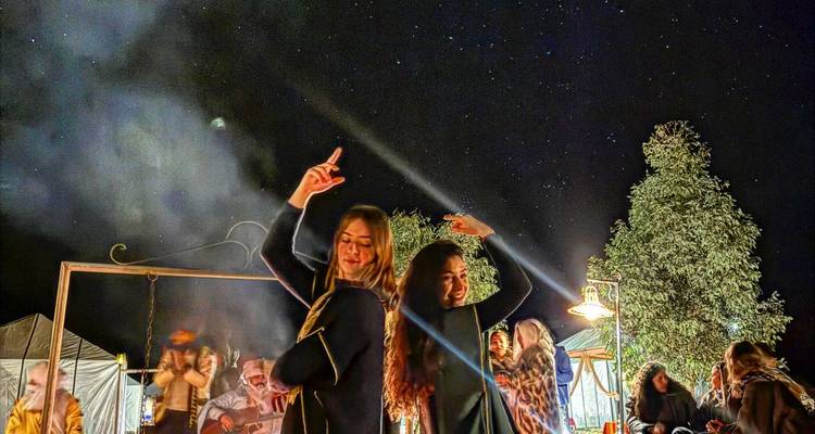 Des voyageurs dansent autour d'un feu de camp sous un ciel nocturne étoilé dans un campement du désert.