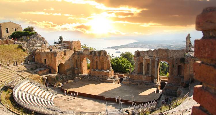 Le théâtre grec antique de Taormine surplombe la mer Ionienne scintillante au coucher du soleil avec des nuages dorés qui remplissent le ciel.