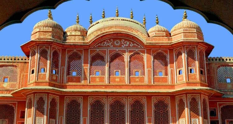 Fachada ornamentada de palacio de arenisca roja con ventanas enrejadas y chhatris abovedados enmarcados por un arco festoneado contra un cielo azul despejado.
