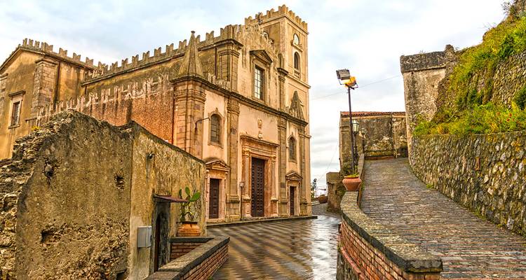 Église en pierre historique et murs de château d'un village sicilien perché sur une colline par temps nuageux.