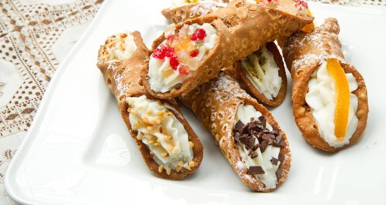 Cannoli siciliens assortis fourrés à la ricotta et décorés de fruits, noix et chocolat sur un plateau blanc.
