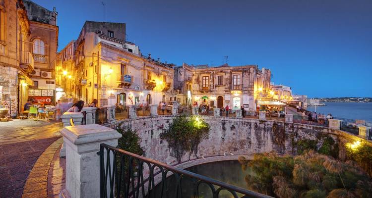 Scène crépusculaire de la place du front de mer de Syracuse avec un éclairage de rue chaleureux se reflétant sur les bâtiments historiques en pierre au bord de la mer.