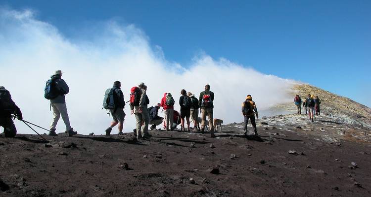 File de randonneurs en équipement de plein air marchant le long du rebord cendreux d'un versant volcanique actif avec de la vapeur qui s'élève.