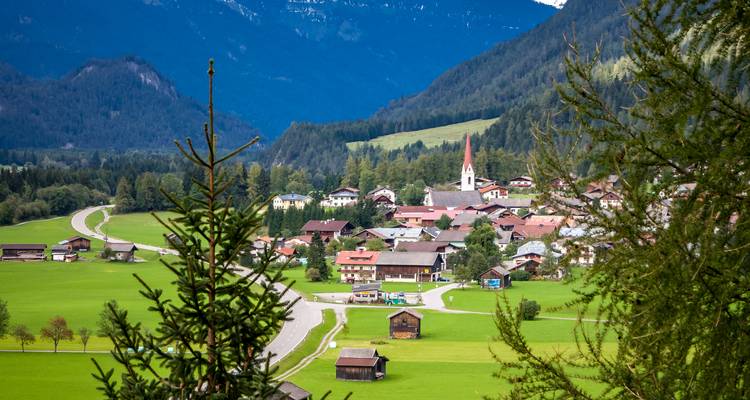 Blick durch Tannenzweige auf ein Taldorf mit Alpendächern und gewundener Straße inmitten üppiger Felder und Berge.