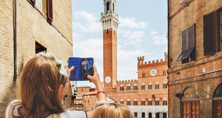Dos chicas fotografiando la Torre del Mangia de Siena entre calles estrechas.