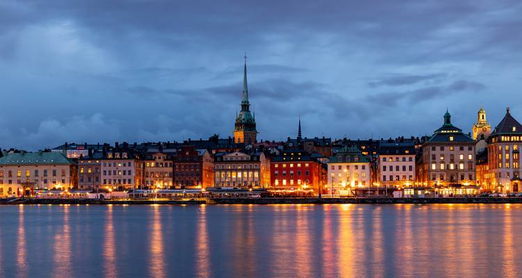 Stockholms Gamla Stan Uferpromenade bei Abenddämmerung mit historischen Gebäuden und Kirchturm, die sich in ruhigem Wasser spiegeln, und Stadtlichtern.