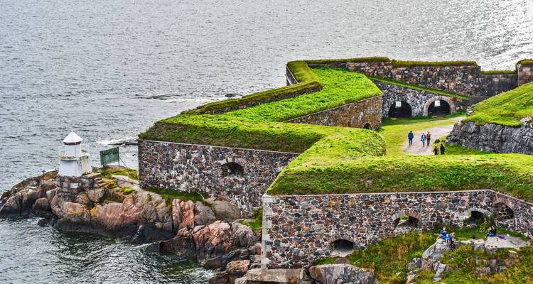 Steinwälle und grasbewachsene Mauern einer Seefestung, die in graue Gewässer hineinragt, mit Besuchern, die entlang des Pfades wandeln.