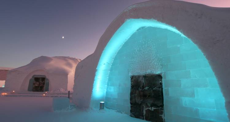 Eisbauwerke, die mit türkisfarbenem Licht gegen eine verschneite Landschaft unter einem blassen Abendhimmel und einer Mondsichel leuchten.