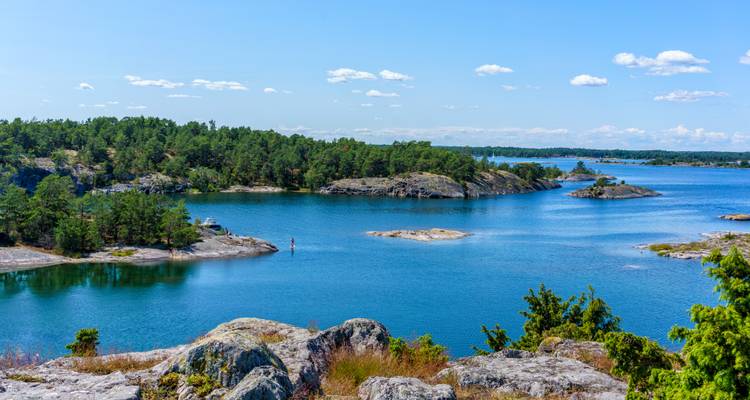 Sonnige schwedische Schärenlandschaft mit kleinen felsigen Inselchen, Kiefern und glitzerndem blauem Wasser, das sich bis zum Horizont erstreckt.