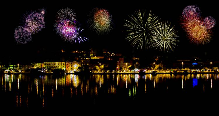 Fuegos artificiales nocturnos estallan sobre una ciudad costera croata, con reflejos brillando en el agua.