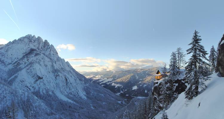 Vallée alpine enneigée avec un pic déchiqueté imposant et une petite cabane illuminée perchée sur une falaise au crépuscule