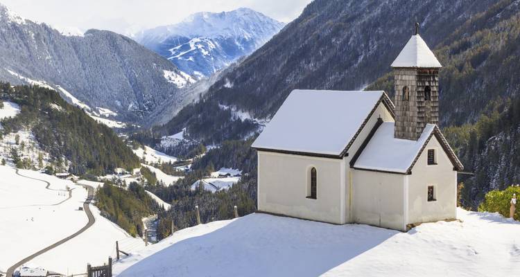 Petite chapelle surmontée de neige surplombant une vallée alpine profonde avec des montagnes bleues au loin