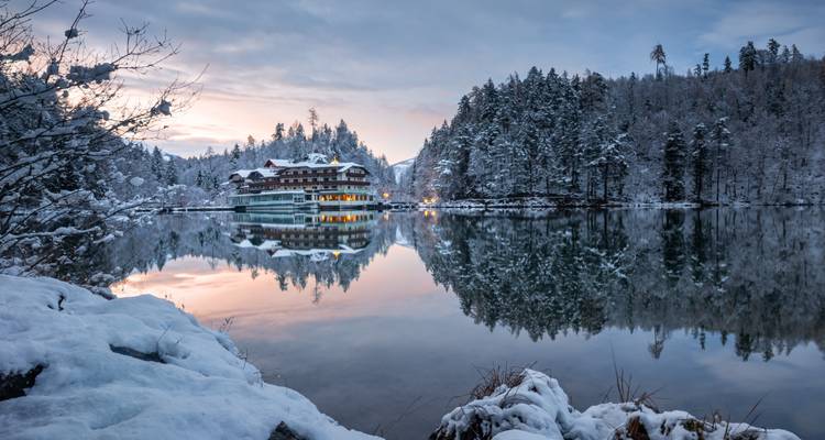 Lac gelé serein reflétant une forêt enneigée et un chalet dans les douces teintes du lever du soleil