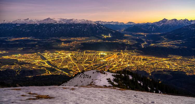 Vue aérienne nocturne sur des montagnes saupoudrées de neige donnant sur les lumières illuminées de la ville d'Innsbruck au crépuscule.