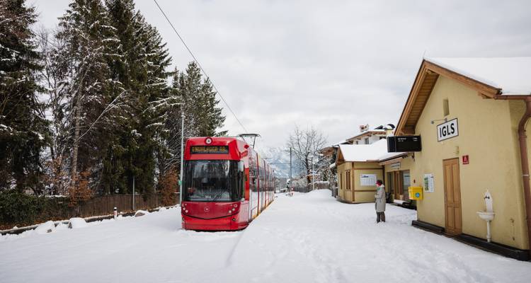 Tramway rouge vif attendant à une petite gare enneigée nommée Igls avec des pins et des montagnes en arrière-plan.