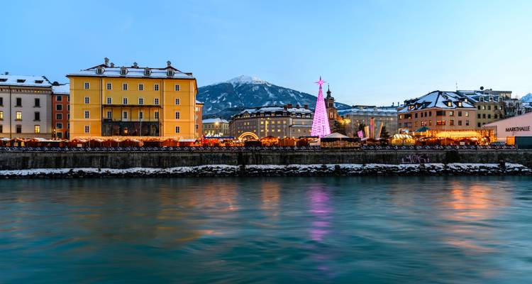 Étals de marché de Noël au bord de la rivière et arbre illuminé à Innsbruck avec des montagnes enneigées en arrière-plan au crépuscule.