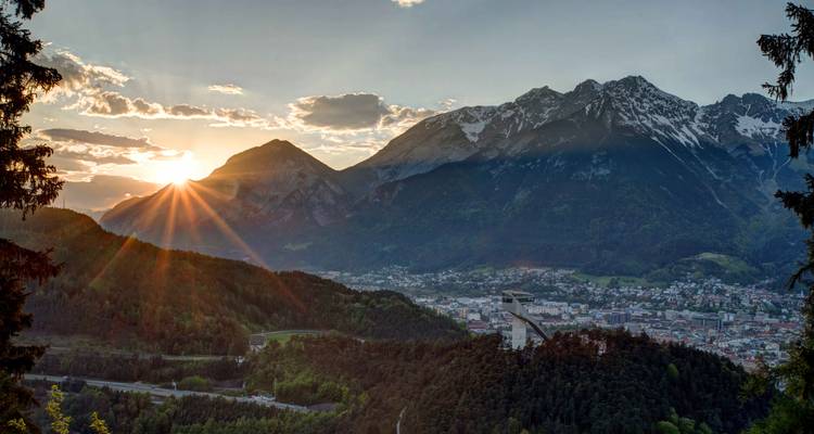 Soleil couchant derrière les sommets alpins avec des rayons se projetant sur une vallée verdoyante et Innsbruck en contrebas.