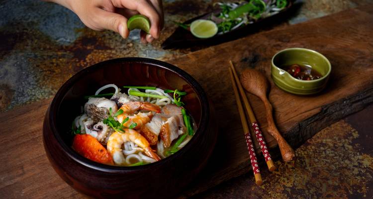 Primer plano de una vibrante sopa vietnamita de fideos con mariscos mientras una mano exprime lima fresca sobre el tazón.